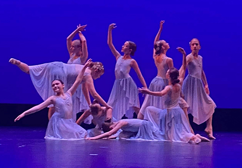 A group of ballet dancers in flowing blue costumes perform a graceful routine on stage, striking elegant poses against a purple-lit background.