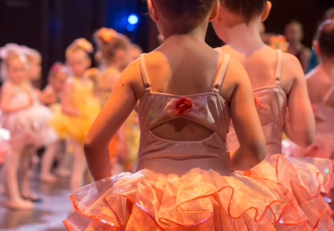 Young children in pastel tutus stand on stage, preparing for a dance recital. The focus is on two dancers in the foreground, with others blurred in the background under warm, colorful stage lighting.