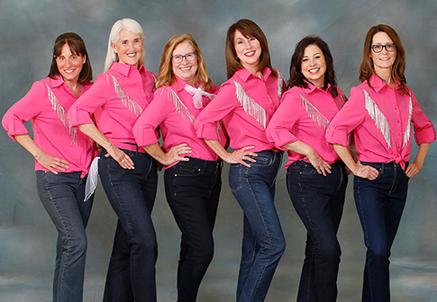 Six women pose in a line, smiling, wearing matching pink shirts with fringes and blue jeans against a gray backdrop. They stand with hands on their hips and lean slightly to one side.