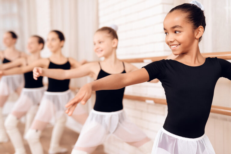 A group of young girls in ballet attire practice at a barre in a dance studio. One girl in the foreground smiles and focuses on her pose, while others in the background follow along.