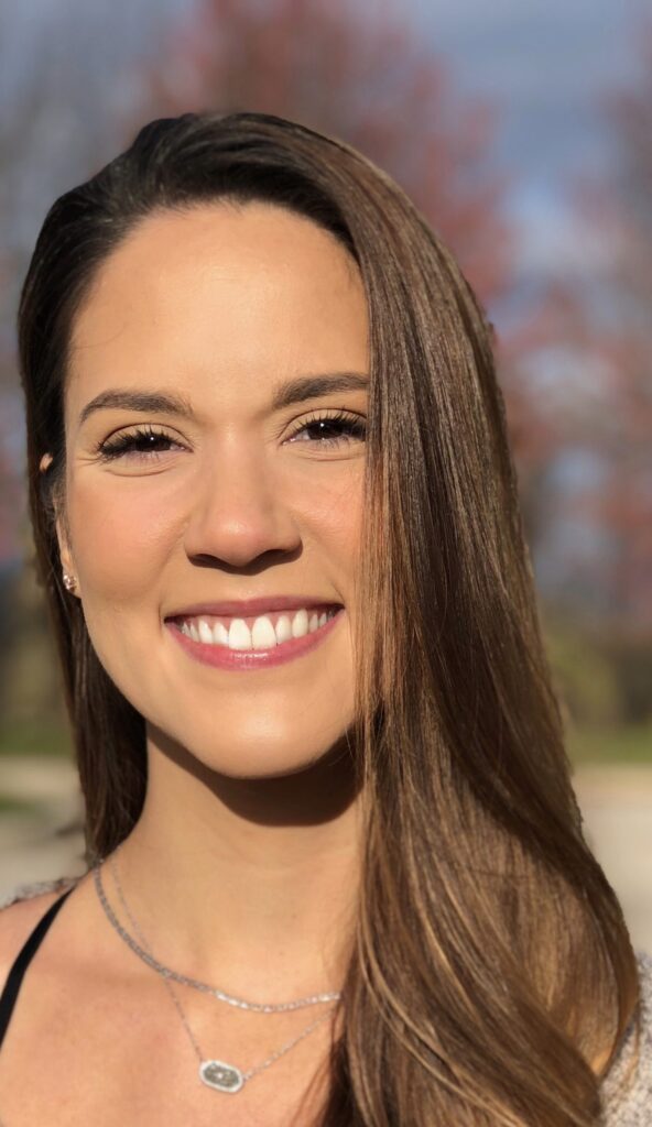 A young woman with long brown hair smiles outdoors on a sunny day. She wears a silver necklace and a light top. The background is blurred, showing trees with reddish leaves and a blue sky.