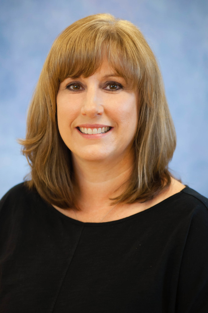 A woman with shoulder-length light brown hair and bangs, wearing a black top, smiles at the camera against a soft blue background.