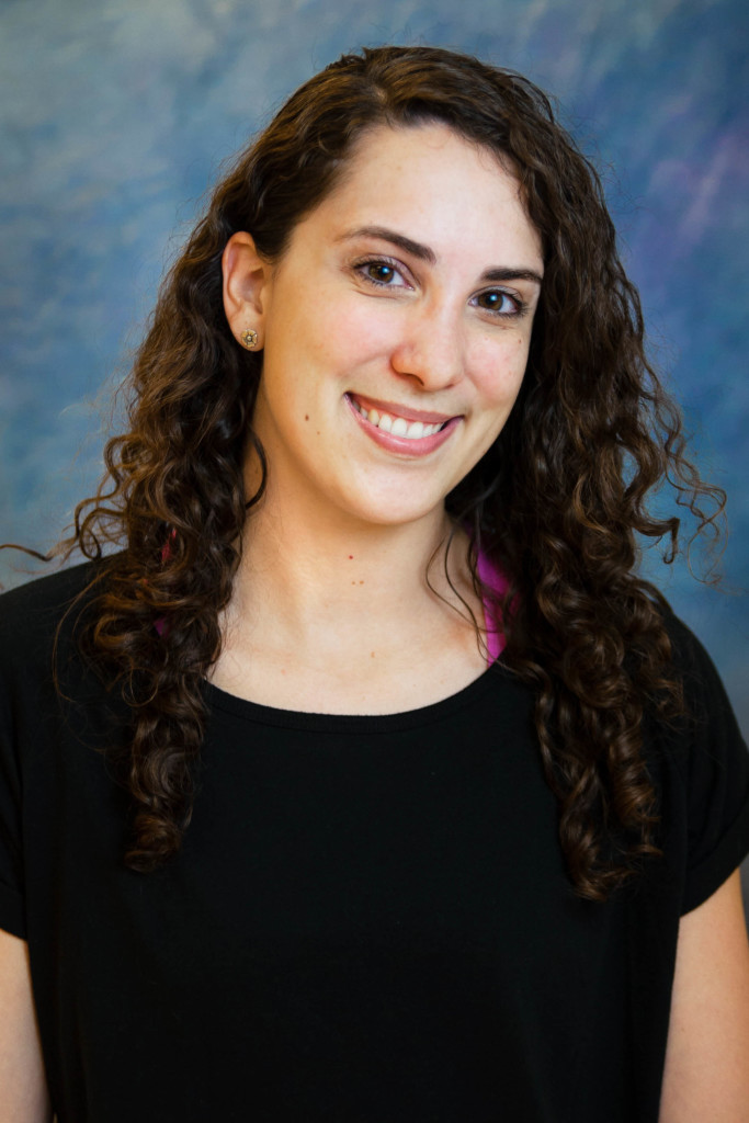 A young woman with long, curly brown hair smiles at the camera. She is wearing a black top and stands in front of a blue, softly blurred background.