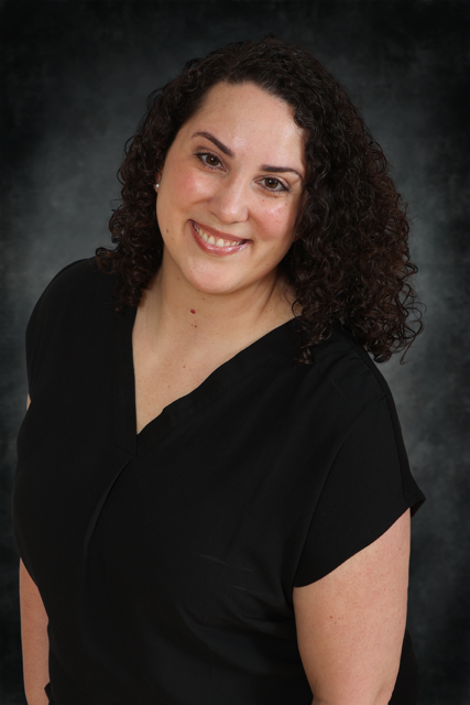 A woman with curly dark hair, wearing a black short-sleeve top, smiles at the camera. The background is dark and subtly textured, creating a professional portrait setting.