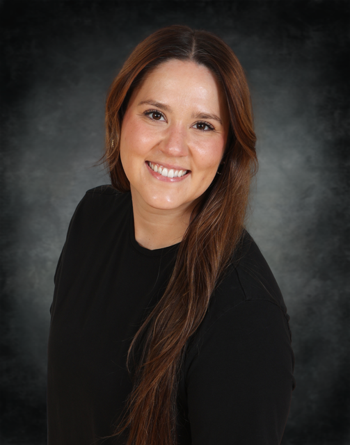 A woman with long brown hair and a black shirt smiles at the camera against a dark, textured background.