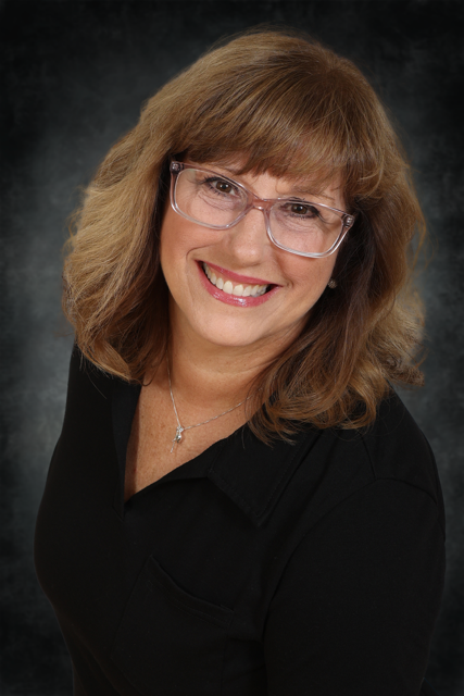 Smiling woman with shoulder-length brown hair, wearing glasses, a black top, and a silver necklace, posed against a dark, textured backdrop.