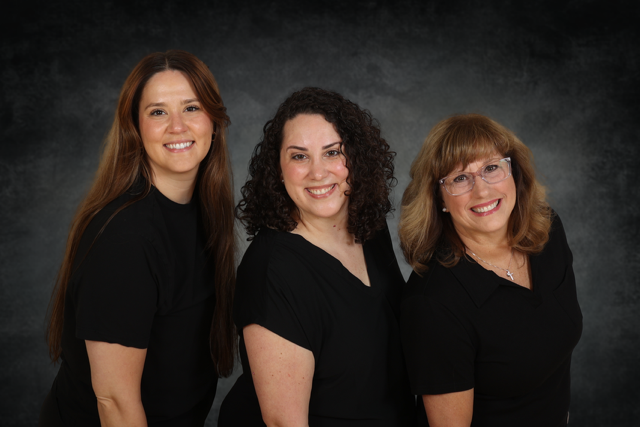 Three women with brown hair pose together against a dark, textured background. All are smiling and wearing black tops.